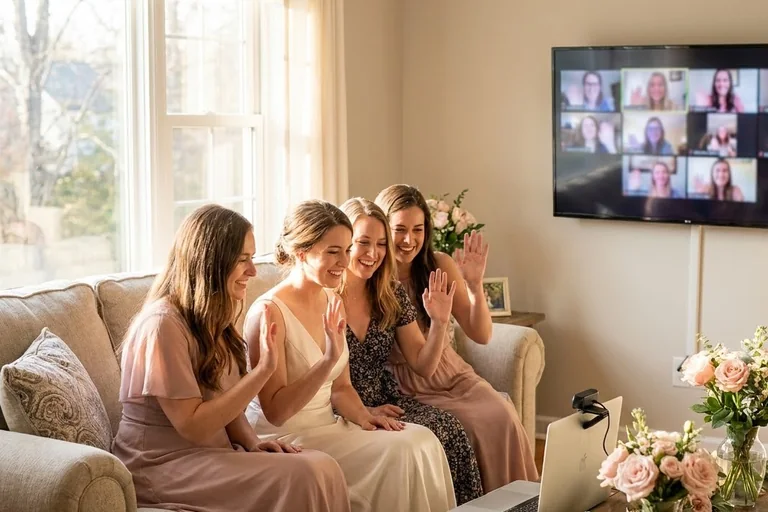 Guests at a bridal shower wave at a TV showing remote friends on a video call while the bride sits beside a laptop and a small microphone.